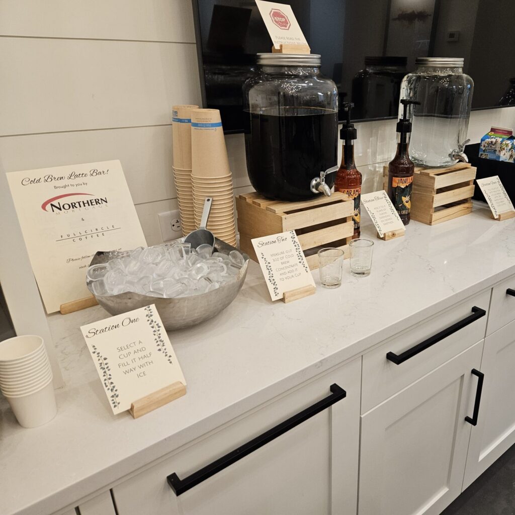 A cold brew latte bar setup in an office, featuring coffee dispensers, flavor syrups, ice, and cups, ready for a customer appreciation event.