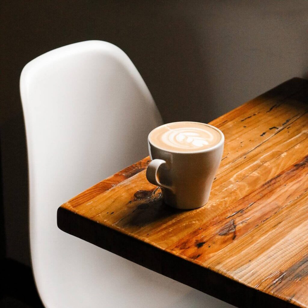 A cappuccino with latte art served in a white mug on a rustic wooden table, next to a white chair in a cozy café setting.