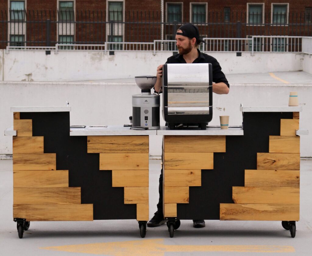 Barista operating a modern wooden and black espresso cart on an outdoor rooftop.